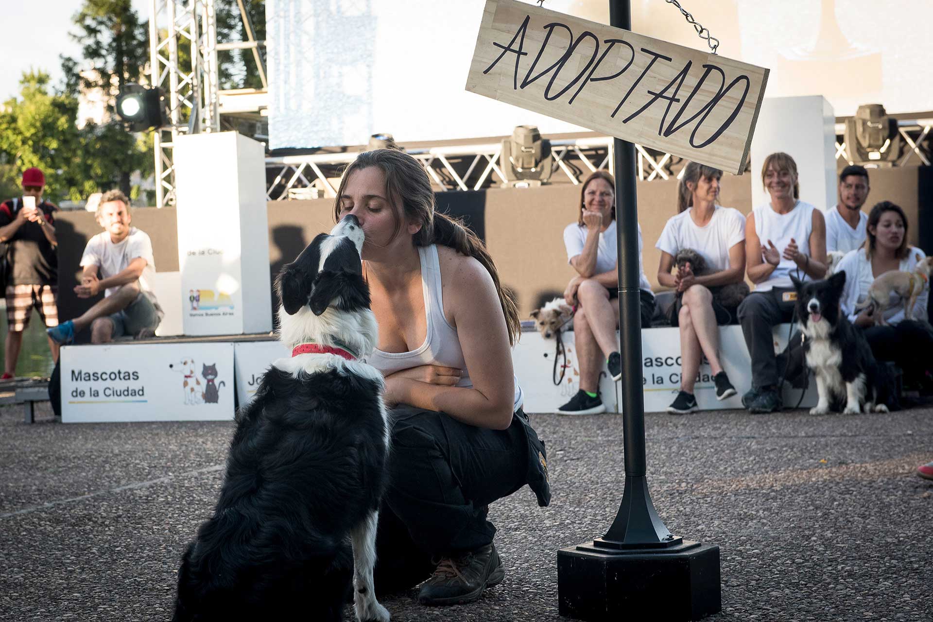Fotografía de una las participantes en cuclillas besando a su mascota en primer plano, de fondo parte de los otros participantes sentados en el escenario. Cobertura del evento 'Mascotas de la Ciudad', organizado por el Gobierno de la Ciudad Autónoma de Buenos Aires