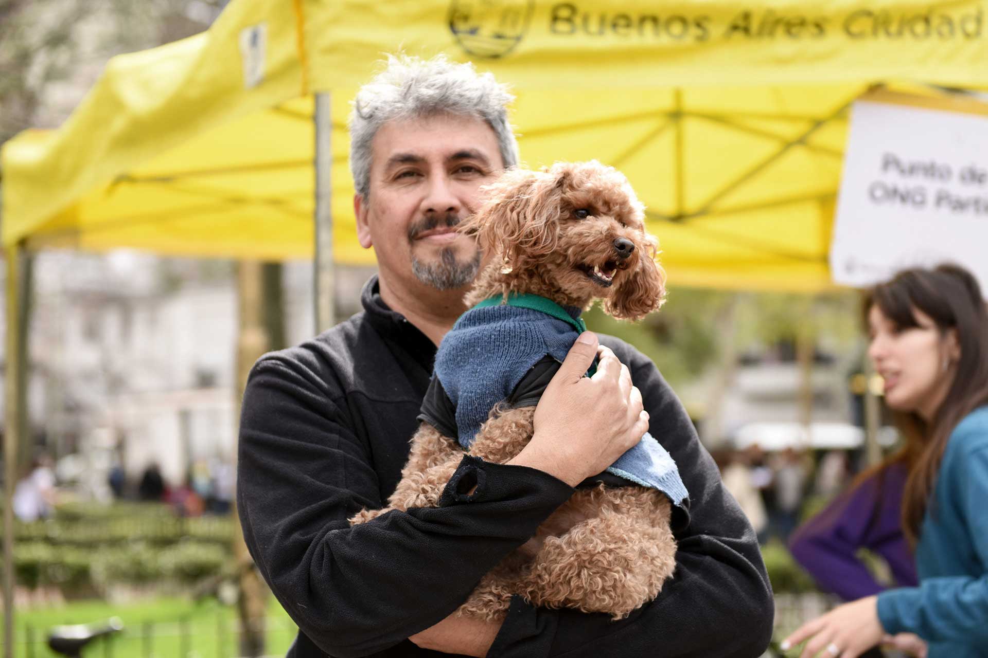 Retrato fotográfico de un hombre con su perro en brazos, la mascota viste un pullover gris oscuro. Cobertura del evento 'Mascotas de la Ciudad', organizado por el Gobierno de la Ciudad Autónoma de Buenos Aires