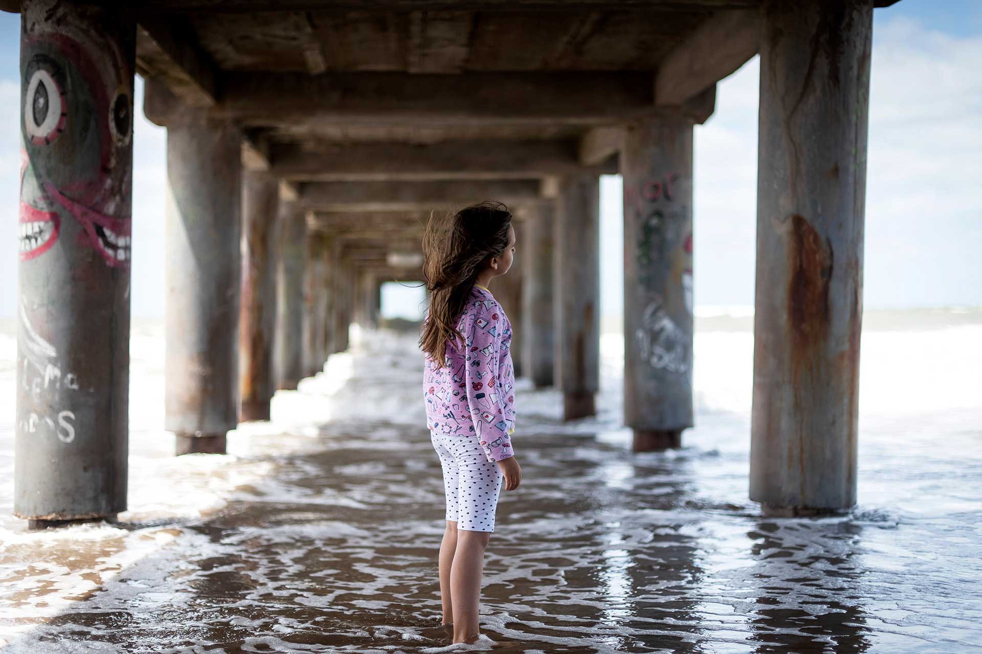 Fotografía de una niña disfrutando del viento en su cara, debajo de un muelle en la playa